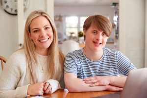 Portrait Of Downs Syndrome Man Sitting With Home Tutor Using Laptop For Lesson At Home